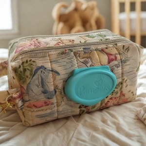 Puede incluir: Un estuche rectangular de tela acolchada con un estampado de Winnie the Pooh y Igor en tonos suaves. El estuche incluye un asa lateral de metal dorado con un cierre de mosquetón y una tapa abatible de plástico color turquesa con el logo Pampers.