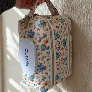 Op de afbeelding: Een luiertas met bloemenprint met een bijpassende doekjeshouder. De tas heeft een crèmekleurige achtergrond met blauwe en roze bloemen en een gouden rits. De doekjeshouder is lichtblauw en heeft het woord "Coterie" erop.