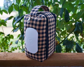May include: A rectangular diaper wipes case with a handle, made of tan and black checkered fabric. A white plastic dispenser is attached to the side. The case is zippered and sits on a wooden surface, with greenery in the background.