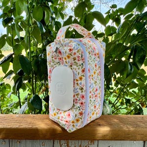 Pode incluir: Uma bolsa de fraldas com estampa floral com um dispensador de lenços umedecidos branco anexado. A bolsa tem uma alça e um fecho de zíper. O fundo é uma superfície de madeira com folhagem verde.