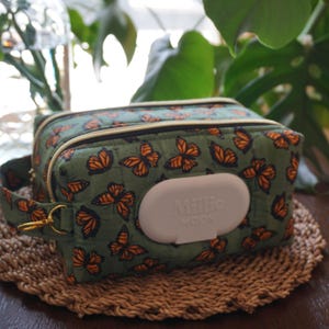 Peut inclure: Une pochette rectangulaire en tissu matelassé vert sauge avec un motif répétitif de papillons monarques. La pochette comprend une poignée latérale avec un anneau en métal doré et un couvercle distributeur en plastique blanc avec le texte Millie Moon.