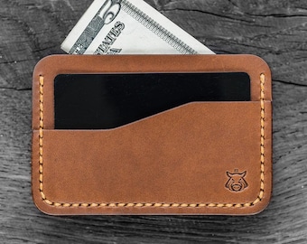 Puede incluir: Una cartera de cuero marrón con una sola ranura para tarjeta y un bolsillo para billetes. La cartera tiene un pequeño logotipo en relieve de una cabeza de jabalí en la esquina inferior derecha.