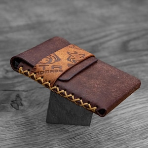 Peut inclure: Un portefeuille en cuir marron avec un bord cousu et une sangle en cuir beige. La sangle porte le texte "STR HANDMADE" et un motif décoratif. Le portefeuille est présenté sur une surface gris foncé.