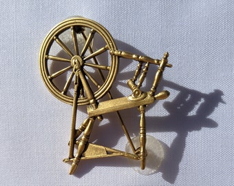May include: A gold-toned spinning wheel brooch with a wooden design. The wheel has spokes and a center hub. The brooch is about 2 inches in diameter.