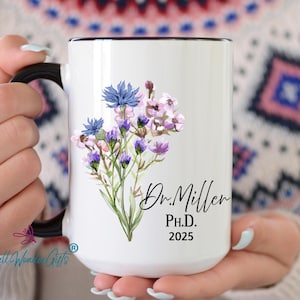 May include: White ceramic mug with a black rim and handle. The mug features a watercolor illustration of purple and blue flowers with green stems. The text "Dr. Miller Ph.D. 2025" is printed in black below the flowers.