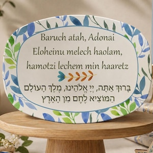 May include: An oval ceramic plate with a floral border, featuring text in English and Hebrew, rests on a wooden cake stand. The plate's design includes blue and green leaves. A lit candle and a Star of David are also visible.