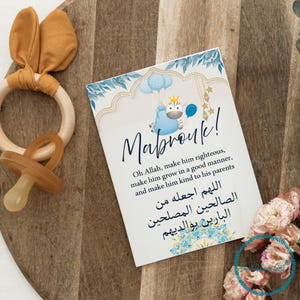May include: A wooden baby teether with a bunny ear design and a pacifier next to a card with the word "Mabrouk!" and a blue and gold design. The card has text in English and Arabic.