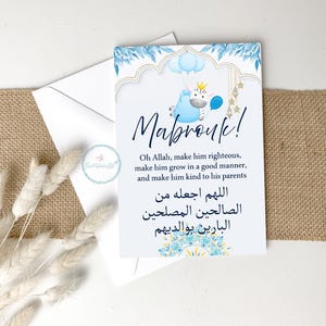 May include: A white card with blue floral and gold accents, featuring the word "Mabrouk!" and a blue cartoon character holding balloons. The card includes text in English and Arabic, and is placed on a burlap surface.