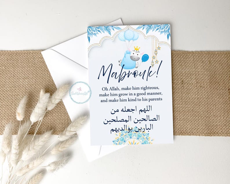 May include: A white card with blue floral and gold accents, featuring the word "Mabrouk!" and a blue cartoon character holding balloons. The card includes text in English and Arabic, and is placed on a burlap surface.