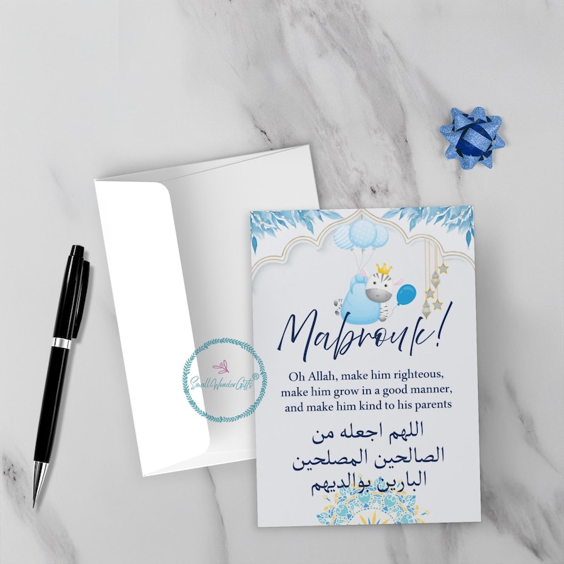 May include: A white greeting card with a blue and gold design, featuring the word "Mabrouk!" and a blue cartoon zebra holding a balloon. The card includes text in English and Arabic, along with a white envelope and a black pen.
