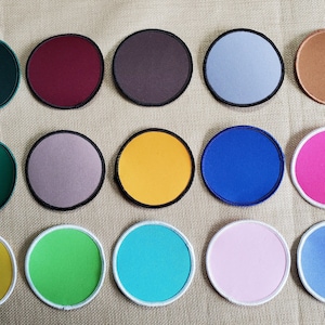 May include: A collection of round, colorful patches with embroidered edges. The patches come in various colors, including green, burgundy, brown, silver, copper, yellow, blue, pink, and turquoise. The patches are arranged in three rows.