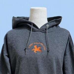 Peut inclure: Un sweat-shirt à capuche gris anthracite foncé avec une poche kangourou sur le devant et des manches longues. La poitrine présente un motif orange brodé représentant un cheval ailé avec le texte "CAMP HALF-BLOOD" arqué au-dessus.
