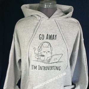 Könnte beinhalten: Ein hellgrauer Kapuzenpullover mit Kordelzugkapuze und Vordertasche. Auf der Vorderseite befindet sich eine schwarz-weiße Grafik einer Katze mit Kopfhörern, die eine Tasse hält und einen Laptop benutzt. Der Text lautet "GO AWAY I'M INTROVERTING" und "EMBROIDERRIFIC.COM".