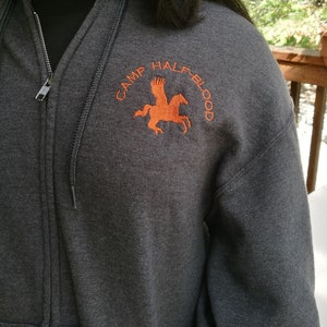 Könnte beinhalten: Ein grauer Kapuzenpullover mit Reißverschluss und einem orangefarbenen gestickten Logo mit der Aufschrift "Camp Half-Blood" und einem geflügelten Pferd.
