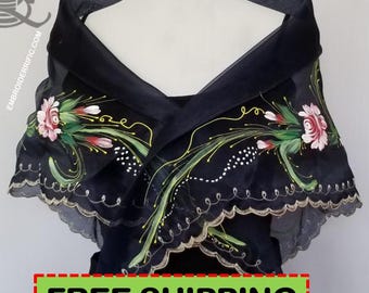 May include: A black sheer shawl featuring hand-painted floral motifs in shades of pink, white, and green. The garment has a wide, folded collar design with scalloped edges finished in a light metallic trim. The fabric is translucent and lightweight.