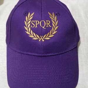 Könnte beinhalten: Lilafarbene Baseballkappe mit einem goldenen, gestickten Lorbeerkranz und dem Text "SPQR" in der Mitte.