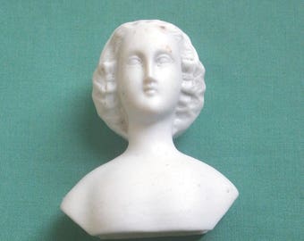 May include: A white porcelain doll head and bust, featuring a serene face with detailed facial features and curly hair. The bust is set against a teal background, highlighting the delicate craftsmanship of the antique doll.