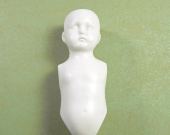 May include: A small, white porcelain doll torso with a rounded head and a pointed lower body. The doll has a smooth, matte finish and is set against a light green background. The doll's face has a slightly sad expression.