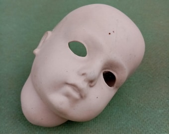 May include: A porcelain doll head with painted features, including eyes, nose, and mouth. The head is unpainted and ready for customization.