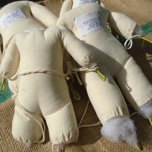 May include: Three beige fabric doll bodies with attached limbs, lying on a burlap surface. Each doll has a label attached. The limbs are secured with string and yellow and green wire. The dolls are stuffed with white material.