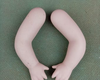 May include: Two porcelain doll arms with hands, in a light pink color. The arms are slightly bent, with fingers partially curled. The doll arms are set against a green background.
