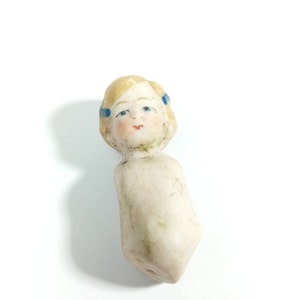 May include: A vintage porcelain doll head with blonde hair and blue painted eyes. The doll has a painted face with a pink mouth and blue ribbons in its hair.