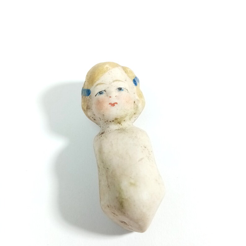 May include: A vintage porcelain doll head with blonde hair and blue painted eyes. The doll has a painted face with a pink mouth and blue ribbons in its hair.
