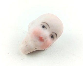 May include: A vintage porcelain doll head with painted features. The doll head has a pale complexion, rosy cheeks, small black eyes, and red lips. The head is bald and appears to be a collectible item.