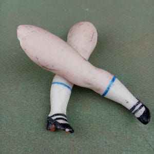 May include: A pair of antique porcelain doll legs, crossed at the ankles. The legs are painted with a pale skin tone, white socks with blue stripes, and black and white shoes with small heels. The legs are resting on a green surface.