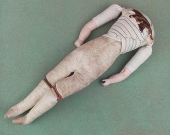 Può includere: Un busto di bambola antica con arti staccati, appoggiato su una superficie verde. Il corpo della bambola è vestito con una camicia a righe e pantaloni beige. Le braccia e le gambe sono di colore rosa pallido e la testa della bambola è mancante.
