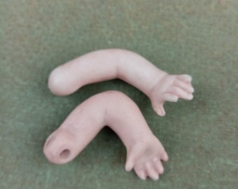 Puede incluir: Dos brazos de muñeca de porcelana con manos, un brazo está doblado en el codo y el otro está recto. Los brazos son de color rosa pálido.