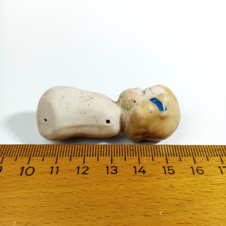 May include: A small porcelain doll figurine with a painted face and blue eyes. The doll is lying on its side with its head turned to the side. The doll is about 4 inches long.