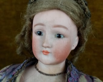 Può includere: Una bambola di porcellana con il viso dipinto e i capelli castani raccolti in uno chignon. La bambola indossa un vestito marrone e viola con un bordo di pizzo.