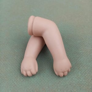 May include: Two porcelain doll arms, painted in a flesh tone, are crossed. The arms are bent at the elbows, with the hands in a slightly clenched position. The upper arm has a cylindrical attachment for the doll's body.
