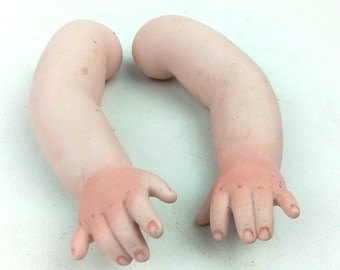 May include: A pair of antique doll arms, made of porcelain. The arms are a pale pink color with darker pink shading on the hands. The fingers are slightly curved, and the nails are detailed.