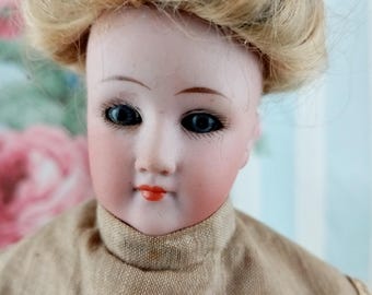 Può includere: Primo piano del viso di una bambola di porcellana vintage. La bambola ha occhi blu, guance rosee e labbra rosse. La bambola ha i capelli biondi e indossa un vestito color beige.