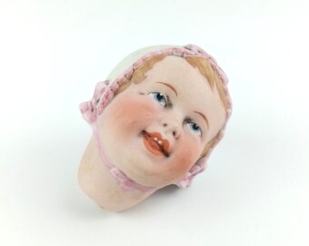 May include: A vintage porcelain doll head with a painted face. The doll head has blue eyes, rosy cheeks, and a small, open mouth. It is wearing a white bonnet with pink trim and a pink ribbon. The doll head is a collectible item.
