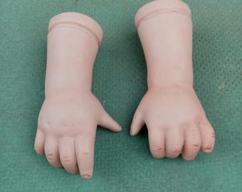 May include: Two antique porcelain doll arms, featuring a pale skin tone and detailed fingers. The arms are slightly bent at the wrists, suggesting a pose. The doll arms are likely vintage and intended for restoration or crafting projects.