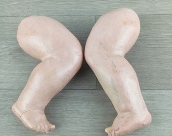 May include: Two pale pink plaster doll legs, bent at the knee, with the feet pointing in opposite directions. The legs are on a light wood surface.
