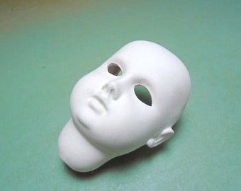 Op de afbeelding: Een witte keramische poppenkop met open oogkassen, een kleine neus en een gedefinieerde kin. De poppenkop is ongeverfd en rust op een lichtgroen oppervlak. Een knutselbenodigdheid.