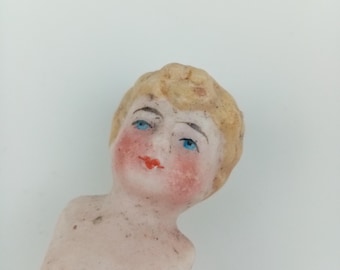 May include: A porcelain doll head and torso featuring molded blonde hair, painted blue eyes, and red lips. The pale skin tone has a matte finish with visible speckling and rosy cheeks. The figure is depicted from the chest up with a neutral expression.