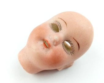 May include: A porcelain doll head with closed eyes and painted eyelashes. The doll head is a light pink color and has a slightly open mouth with a painted red lip.