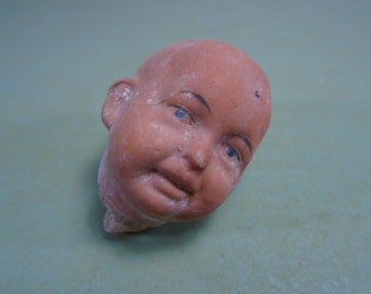 May include: A terracotta doll head with painted eyes and a closed mouth. The doll head is slightly tilted to the side.