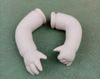 Może przedstawiać: Dwa odłączone, białawe porcelanowe ramiona lalki. Ramiona są zgięte w łokciu, z lekko zakrzywionymi dłońmi. Ramiona są umieszczone na zielonym tle. Ramiona są prawdopodobnie częściami zamiennymi do zabytkowej lalki.