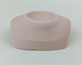 May include: A small, light pink ceramic candle holder with a circular opening. The candle holder has a rounded, square shape with a smooth surface. The opening is centered on the top, designed to hold a small candle.