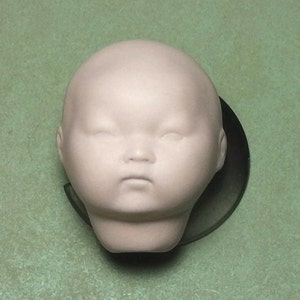 May include: A white porcelain doll head with closed eyes and a neutral expression. The head is smooth and has a slightly rounded shape.