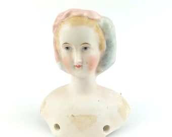 May include: A porcelain doll head with a pink and blue bonnet. The doll has painted blonde hair, blue eyes, and rosy cheeks.