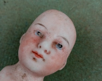 May include: A porcelain doll head with painted blue eyes, rosy cheeks, and a closed mouth. The doll's head is slightly tilted to the side.