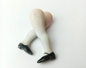 May include: A pair of porcelain doll legs with white stockings and black shoes. The legs are posed in a standing position with one leg bent at the knee.
