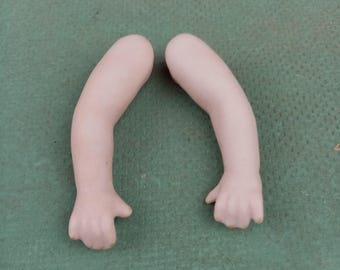 Może przedstawiać: Dwa porcelanowe ramiona lalki z dłońmi, w jasnym odcieniu skóry. Ramiona są zakrzywione, z dłońmi lekko otwartymi. Tło to jednolita, teksturowana, ciemnozielona powierzchnia.